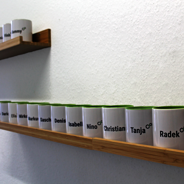 Foto: Kaffeebecher mit Namen der Mitarbeiter stehen auf einem Wandregal