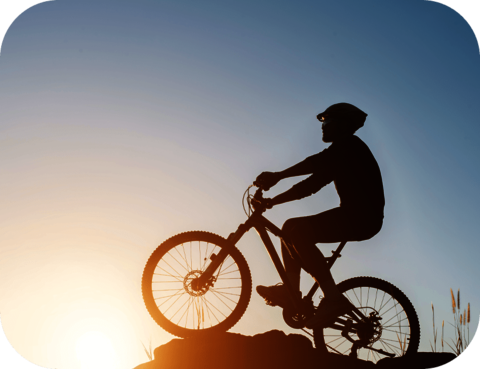 Foto: Silhouette eines Radfahrers mit Helm auf dem Gipfel eines Berges, blauer Himmel mit untergehender Sonne