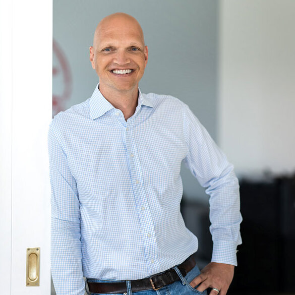 atlantis dx - Denis Rathig, Bereichsleiter E-Commerce Foto: Frontalaufnahme von Denis Rathig, trägt ein weißes Hemd, lehnt entspannt in einem Türrahmen, lächelt freundlich