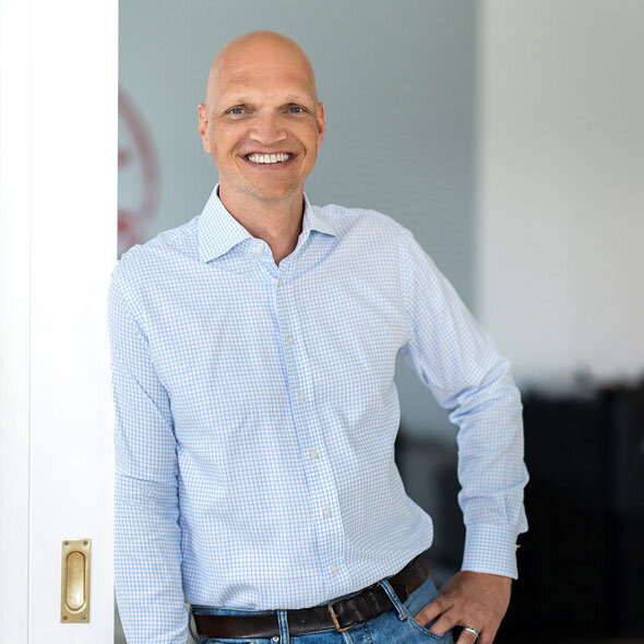 atlantis dx - Denis Rathig, Bereichsleiter E-Commerce Foto: Frontalaufnahme von Denis Rathig, trägt ein weißes Hemd, lehnt entspannt in einem Türrahmen, lächelt freundlich