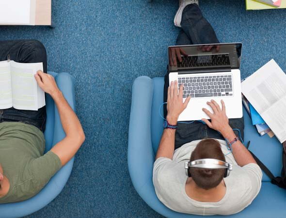 Foto: Sicht von oben auf zwei Studierende, die Laptops auf dem Schoß haben, sitzen locker in blauen Sesseln