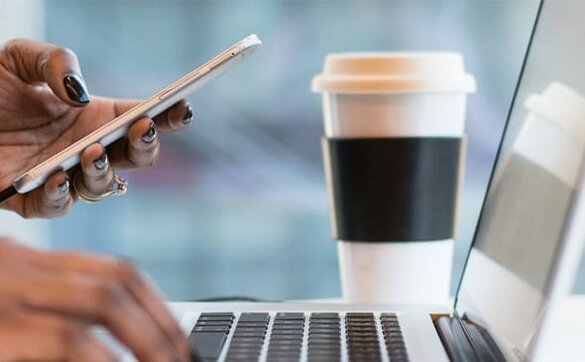 Karriere Foto: Seitenansicht eines geöffneten Laptops, eine Hand auf Tastatur, die andere Hand hält ein Handy, daneben steht ein Coffee-Cup