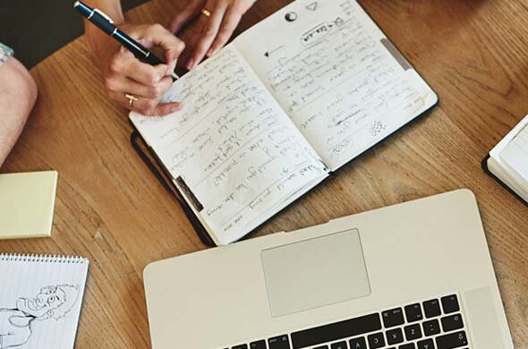 Foto: Sicht von oben auf einen Holztisch mit Laptop, einem Notizbuch in das gerade geschrieben wird, Post It´s, Skizzenblock. Lernsituation