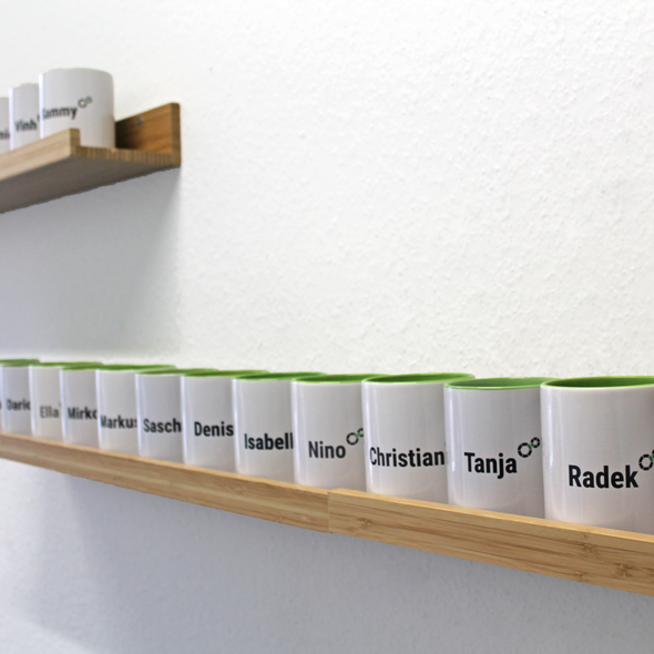 Foto: Kaffeebecher mit Namen der Mitarbeiter stehen auf einem Wandregal