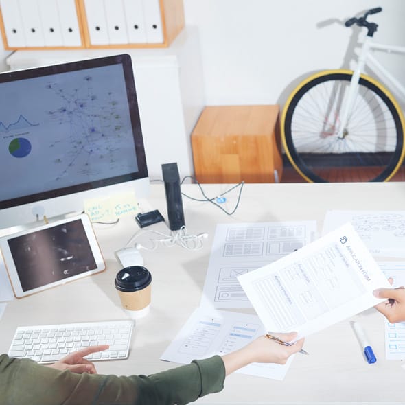Sicht auf weißen Schreibtisch mit Unterlagen, Tablet und Monitor mit Darstellung verschiedener Diagramme, im Hintergrund lehnt ein Fahrrad an der Wand