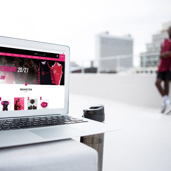 Foto: Outdooraufnahme, Laptop mit geöffnetem Shop von EVENTIM SPORTS, im Hintergrund ein Sportplatz ein Basketballspieler, ein weißes Hochhaus