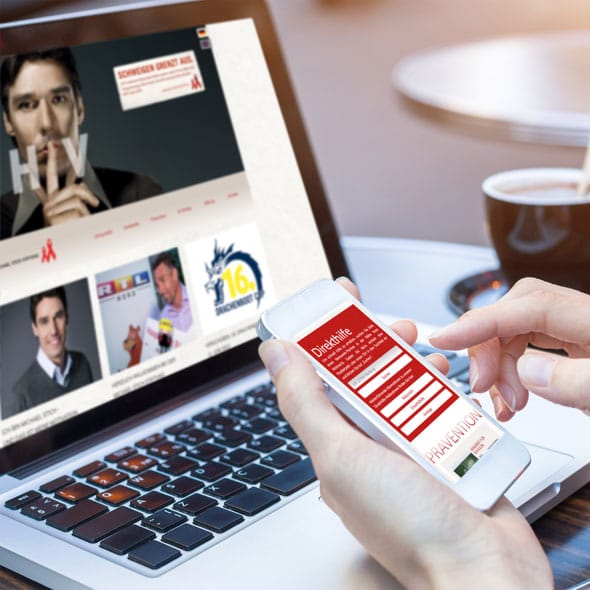 Nahmaufnahme eines Laptops mit geöffneter Website, im Vordergrund hält eine Person ein Handy in der Hand, im Hintergrund eine Tasse Kaffee
